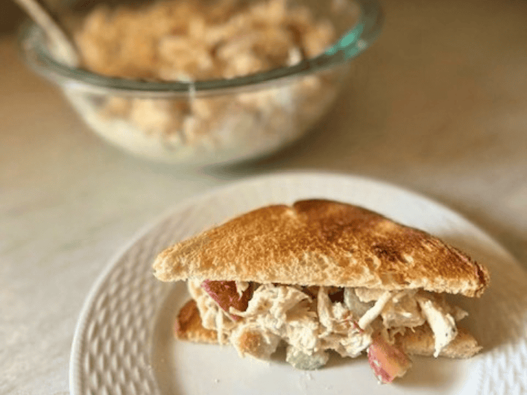 Chicken salad sandwich on a white plate on the kitchen counter with a bowl of chicken salad beside it.
