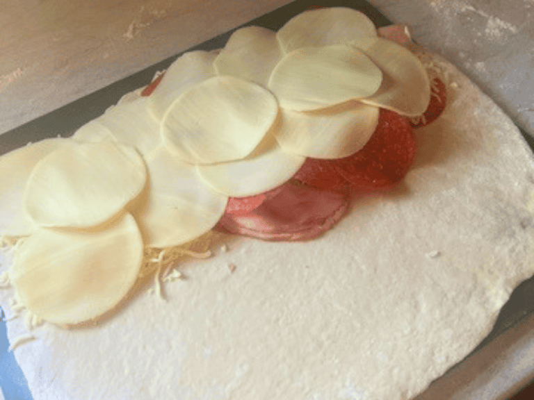 Rolled-out stromboli dough topped with layers of deli meats, mushrooms, and shredded cheese on a baking pan