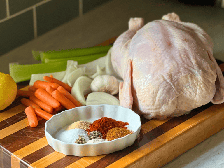Raw whole chicken on a cutting board surrounded by seasoning spices, celery, carrots, onion, and garlic before cooking.