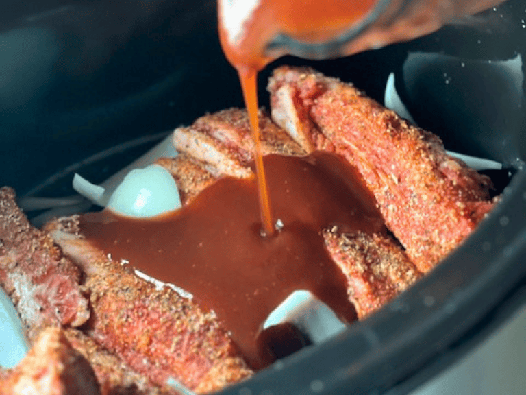Raw beef short ribs layered in a slow cooker with sliced onions, minced garlic, barbecue sauce ingredients, and seasonings before cooking.