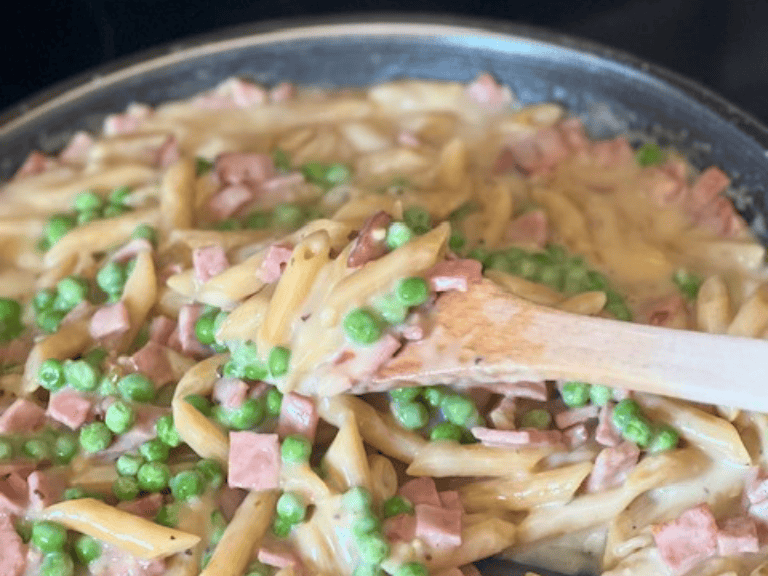 Creamy ham and penne pasta with peas in a skillet on the stovetop, with a wooden spoon resting in the dish.