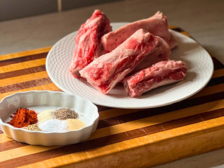 Raw beef short ribs on a cutting board with a small bowl of spices and seasonings prepared for cooking.