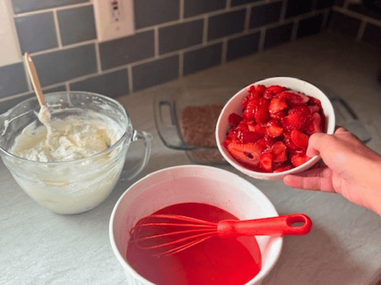 Fresh sliced strawberries being poured from a white bowl into strawberry gelatin in a large glass mixing bowl for Strawberry Pretzel Salad.