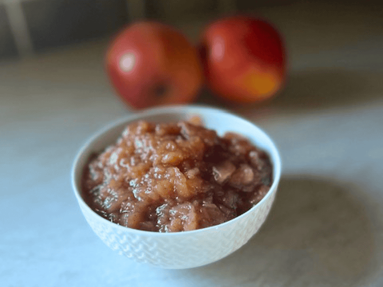 A bowl of homemade slow cooker applesauce with fresh apples on the kitchen counter in the background.