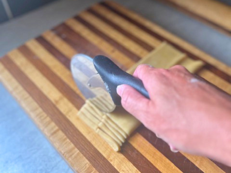 Cutting folded homemade pasta dough into fettuccine strips with a pizza cutter.